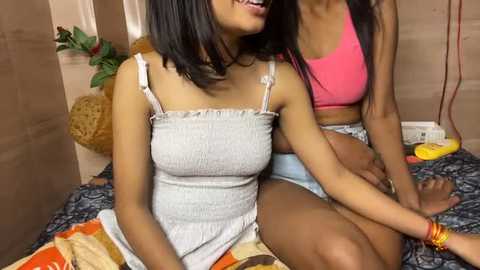 Media: Video of two South Asian women, one in a gray ruffled dress, the other in a pink crop top, sitting on a patterned bedspread.