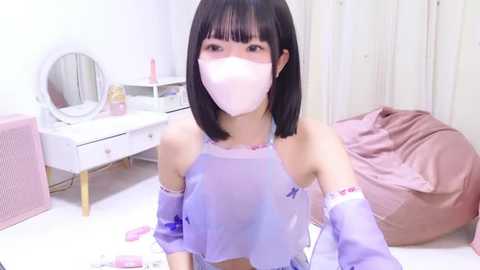 Media: A young Asian woman with straight black hair and bangs, wearing a white mask and a see-through lavender crop top, sits on a bed with a pink blanket. The room features a vanity with a mirror, pink chair, and a soft pink rug.