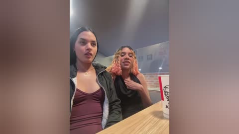 Media: Video of two young women, one with dark hair and the other with orange dreadlocks, standing in a fast-food restaurant. They wear casual clothing, one in a maroon top, the other in a black shirt.