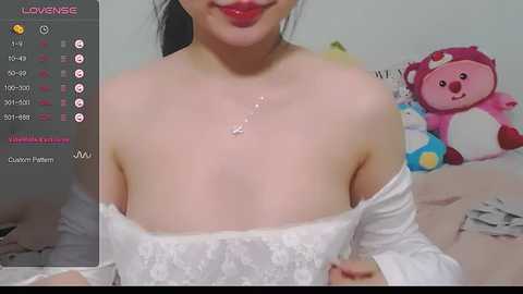 Media: Video of an East Asian woman with fair skin, wearing a white lace off-shoulder top, red lipstick, and a pendant necklace. Background features a plush toy and a gray pillow.