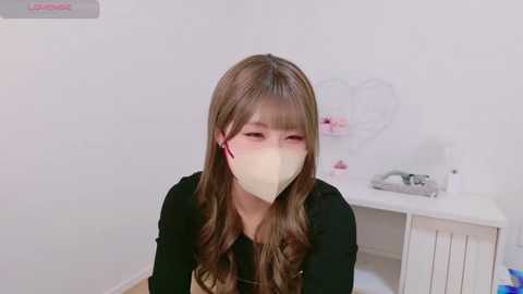 Media: A video of a young Asian woman with long brown hair, wearing a black top and a white mask, sitting in a minimalistic room with white walls and a white desk.