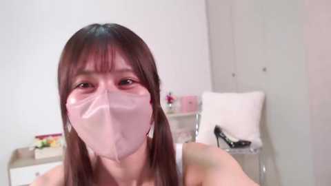 Media: Video of an Asian woman with straight brown hair, wearing a pink face mask and a white top, sitting in a minimalist room with white walls, a chair, and a table.