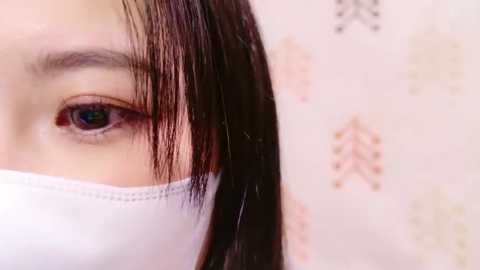 Media: Close-up video of an Asian woman with dark brown hair, partially obscured by a white surgical mask, revealing her left eye with a detailed iris. The background features a beige wall with faint floral patterns.