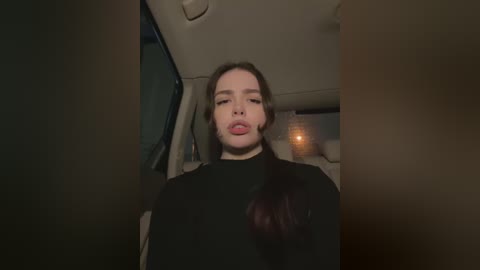 Media: Video of a young Caucasian woman with light skin and long brown hair, wearing a black turtleneck, sticking out her tongue inside a dimly lit car.