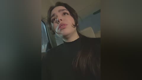 Media: Video of a young woman with light skin and dark hair, wearing a black top, captured from a low angle inside a dimly lit room.