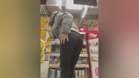 Media: A video of a woman in a grocery store, wearing a grey hoodie and black leggings, bending over, revealing her buttocks. The background features shelves with various products, including a red sign and yellow packages.