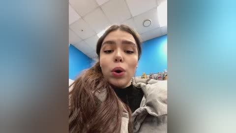 Media: Video of a young woman with long brown hair, light skin, and brown eyes, wearing a beige jacket, in a room with blue walls and white ceiling tiles, making a kissing gesture.