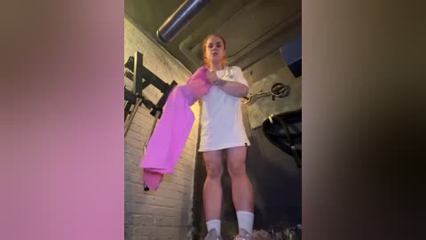 Media: A video of a blonde woman in a white t-shirt and shorts standing in a dimly lit, industrial space with a brick wall and exposed pipes, holding a bright pink jacket.