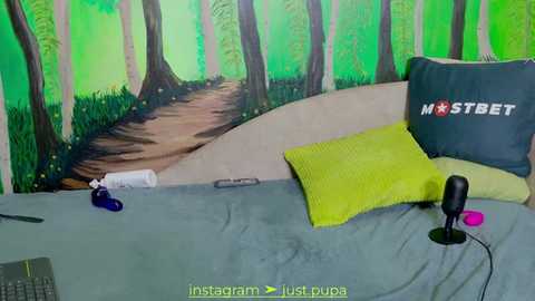 Media: A video of a cozy bedroom featuring a bed with a lime green pillow, a grey pillow with \"MOSTBET\" text, a grey blanket, a black microphone, and a green wall mural depicting a forest path.