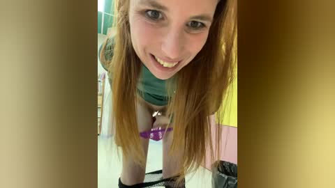 Media: Video of a young woman with long, straight, reddish-brown hair, wearing a green top, bending over, pulling down her purple panties, revealing a purple dildo. Background includes a yellow wall and a pink shelf.