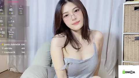 Media: A video of an Asian woman with fair skin, long straight hair, and glasses, wearing a gray tank top, sitting on a bed with a white curtain background.