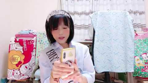 Media: Video of an Asian woman with short black hair, wearing a white headband and a blue shirt, taking a selfie with a yellow phone in a colorful, cluttered bedroom.