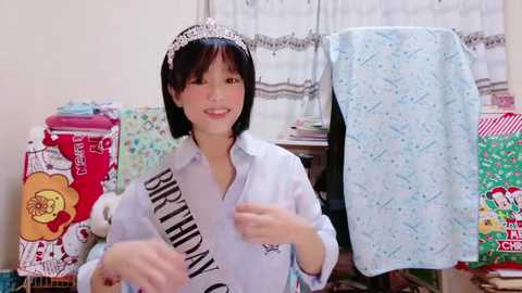 Media: Video of a cheerful Asian woman with short black hair wearing a silver tiara, a \"BIRTHDAY\" sash, and a light blue shirt, surrounded by colorful, patterned blankets and pillows in a cozy, cluttered room.