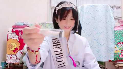 Media: Video of a young woman with straight black hair, wearing a tiara, white shirt with \"Birthday\" sash, and bracelets, holding a pink cake, surrounded by colorful gift boxes in a bright, cluttered room.