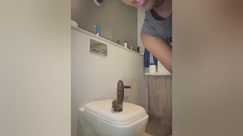 Media: A video of a bathroom with a white toilet featuring a large, realistic brown dildo on the lid. The setting is a modern, minimalist bathroom with beige walls and a white sink. A person, only partially visible, is standing in the background.