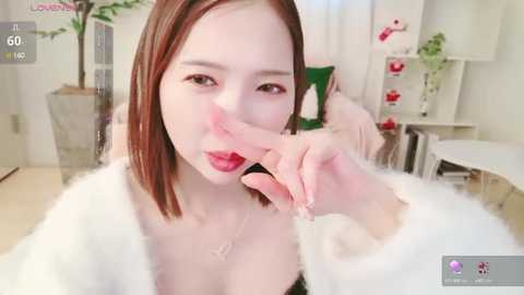 Media: A video of an East Asian woman with fair skin, red hair, and light makeup, wearing a white fluffy robe, playfully covering her mouth with her finger. Background includes plants and a white chair.