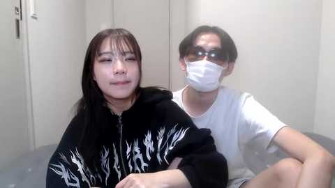 Media: Video of two women, one with long black hair in a black jacket, the other with short black hair and a white mask, seated in a plain, beige room.