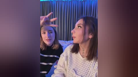 Media: Video of two young girls, one blonde in a striped sweater, the other brunette in a polka-dot top, looking at a TV screen. Background features blue curtains.