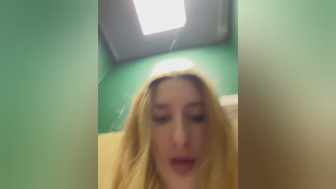 Media: Video of a blonde woman with fair skin and full lips, blurred and slightly out of focus, standing in a green-walled restroom with a yellow towel and a fluorescent ceiling light.