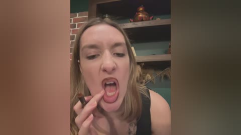 Media: Video of a blonde woman with fair skin and medium build, wearing a black top, playfully sticking out her tongue and touching her lips with her fingers, in a rustic room with wooden shelves and a brick wall.