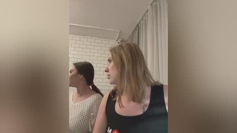 Media: Video of two women in a bathroom; one with light brown hair and a tattoo, the other with dark hair in a ponytail. White tiled walls, beige curtain.