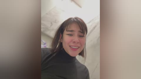 Media: A video of a smiling young woman with straight brown hair, wearing a black turtleneck, standing in a dimly lit bathroom with tiled walls.