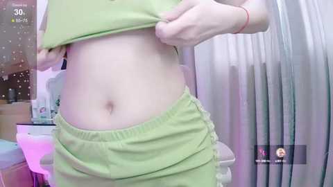 Media: Video of a fair-skinned person lifting a light green top, revealing a flat stomach. They wear matching light green shorts. Background shows a bathroom with a shower curtain, a small table, and a thermometer.