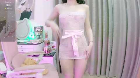 Media: A video of a slender, light-skinned woman in a sheer, pink lace dress, standing in a dimly lit, cluttered bedroom with pastel-colored walls and a vanity table.