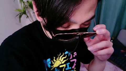 Media: A video of an Asian man with short black hair, wearing a black T-shirt with a colorful sun design, adjusting dark sunglasses, in a room with green curtains and a potted plant.