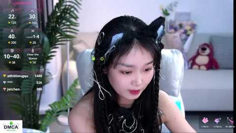 Media: Video of a young East Asian woman with long black hair and cat ears, wearing a black top, sitting in a modern living room with a white couch and green plants.