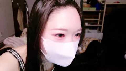 Media: A video of an East Asian woman with long black hair, wearing a white surgical mask and a black top, in a cluttered bedroom with bookshelves and clothes.