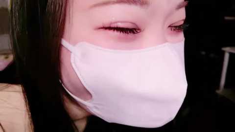 Media: Close-up video of a young Asian woman with long black hair, wearing a white surgical mask, eyes closed, expression serene.