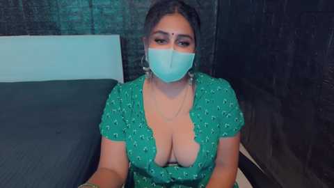 Media: Video of a South Asian woman with medium skin tone, wearing a green floral dress, large earrings, and a teal face mask, sitting on a dark bed with a teal headboard in a dimly lit room.