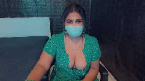 Media: Video of a curvy, light-skinned woman with dark hair, wearing a green dress with a deep V-neck, revealing cleavage, and a teal face mask, seated on a bed with dark sheets and a brick-patterned wall in the background.