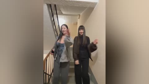 Media: Video of two Asian women in a beige hallway. One wears a gray jacket over a blue dress, while the other has long black hair and is in a black outfit.