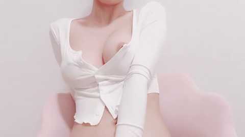 Media: Video of a light-skinned woman with large breasts, wearing a tight, white, long-sleeved crop top with a deep V-neck, revealing cleavage. She sits on a pink chair against a plain white background.