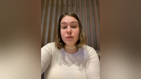 Media: Video of a young Caucasian woman with straight, shoulder-length brown hair, wearing a white long-sleeve shirt and a pearl necklace, standing in front of vertical blinds.