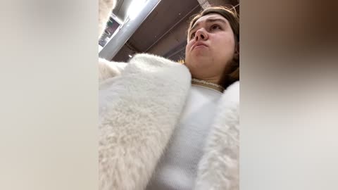 Media: A video shows a young Caucasian woman with shoulder-length brown hair, wearing a white fluffy sweater, looking up with a pensive expression. The background is blurred, featuring a ceiling and fluorescent lights.