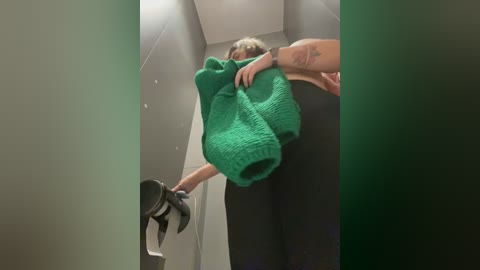 Media: A video captures a woman in a green sweater and black pants, holding a black bag, in an elevator. The background is dimly lit, with gray walls and a visible hand with a tattoo on it.