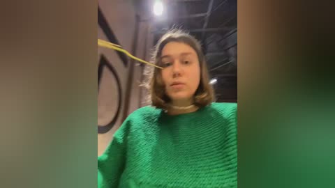 Media: Video of a young woman with shoulder-length brown hair, wearing a green knitted sweater, and a yellow tape measuring her eye. The background features a wooden ceiling and a round clock.