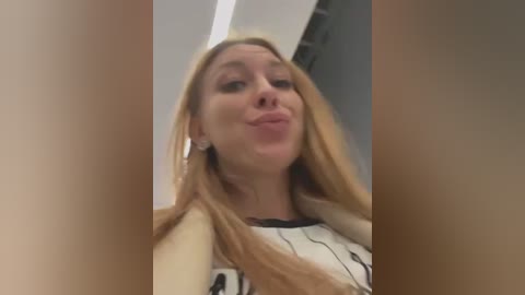 Media: Video of a Caucasian woman with long, straight blonde hair, wearing a white blouse with black abstract designs. She has a playful expression, puckering her lips. Background shows indoor lighting and a blurry, gray wall.