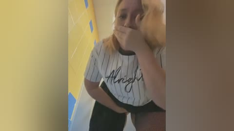 Media: Video of a blonde woman with fair skin, wearing a white pinstriped crop top and black shorts, covering her mouth with her hand, standing in a yellow-tiled bathroom.