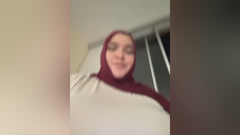 Video of a smiling woman in a maroon hijab, wearing a beige long-sleeve shirt, standing indoors with blinds in the background.