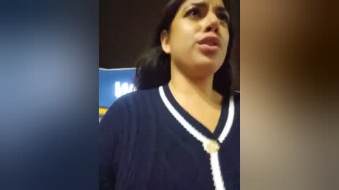 Media: Video of a woman with medium skin tone, dark hair, and a serious expression, wearing a navy blue cardigan with white trim, standing indoors against a blurred background.