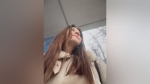 Video of a young woman with long brown hair, wearing a beige coat, tilted head back, smiling, standing in a brightly lit room with a white ceiling and a blurred background.