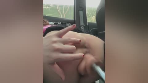 Media: Video of a woman with fair skin, red-painted nails, and a pink top, sitting in a car. She is spreading her labia with fingers, revealing her vulva. The background shows a blurry landscape outside the window.