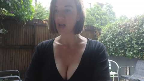 Media: Video of a fair-skinned, short-haired woman with a deep V-neck black top, speaking outdoors in a sunlit garden with greenery and a wooden fence.