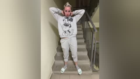 Media: Video of a blonde woman with a slim build, wearing a Mickey Mouse sweatshirt and gray sweatpants, standing confidently with arms raised on a stairwell with beige walls.