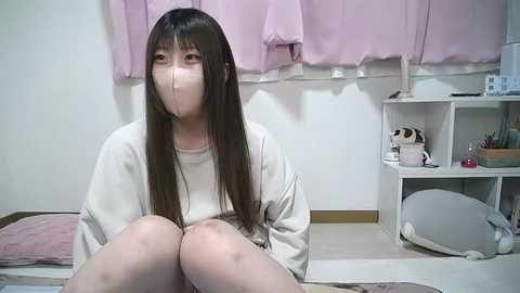 Media: A video of an Asian woman with long, straight black hair, wearing a white mask and oversized sweater, seated on a white cushioned bench in a minimalist room with light pink curtains, shelves with toys, and a gray cushion.