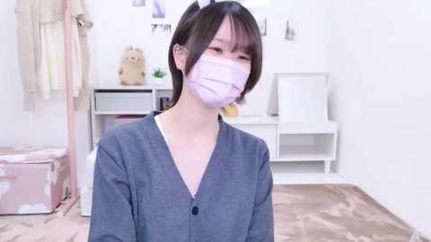 Media: A video of an East Asian woman with short black hair, wearing a gray cardigan, a white mask, and a light pink face shield, standing in a minimalist, white-walled room with a wooden floor.